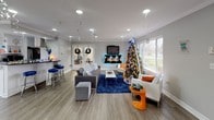This single-floor clubhouse is a thoughtfully designed community space that seamlessly blends functionality with a warm, inviting atmosphere. The main lounge area serves as the heart of the clubhouse, featuring wood-look flooring, crown molding, and recessed lighting throughout. A wall-mounted flat-screen TV is positioned above a fireplace mantle, flanked by comfortable seating arrangements including a plush gray sofa, accent chairs, and a bold blue area rug that anchors the space. Large windows flood the room with natural light while offering pleasant views of the surrounding greenery outdoors. A bar-height seating area with vibrant blue stools provides an additional social gathering spot along one wall. Adjacent to the lounge, a fully equipped kitchen offers ample cabinetry, granite countertops, stainless steel appliances, and a central island. A bistro-style dining table with bar-height chairs sits near sliding glass doors that open to an outdoor patio, enhancing the connection between interior and exterior spaces. A welcoming reception nook features a small counter with a coffee station and a chalkboard, directing residents toward a private office area equipped for administrative needs. The cohesive gray-and-white color palette, crown molding details, and consistent flooring throughout create a polished and unified aesthetic across all areas of this versatile clubhouse.