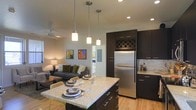This single-floor space showcases a thoughtfully designed open-concept layout that seamlessly connects the kitchen and living areas. The kitchen is a standout feature, boasting rich dark cabinetry with sleek silver hardware, a stainless steel refrigerator, and a marble-style tile backsplash. A large granite-topped island with seating serves as both a functional prep area and an informal dining space, illuminated overhead by a trio of modern pendant lights. A built-in wine rack adds a refined touch to the upper cabinetry. Light hardwood flooring runs throughout, enhancing the warm, contemporary feel of the space. Recessed lighting and a ceiling fan provide practical comfort across the open floor plan. The living area flows naturally from the kitchen, furnished with a comfortable sofa accented with colorful throw pillows and complemented by warm ambient lighting from a classic table lamp. Colorful wall art adds personality and visual interest to the neutral-toned walls. A glimpse into an adjoining room suggests additional private living space beyond. Overall, the space strikes a balance between modern design and comfortable, everyday livability.