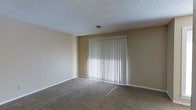 This single-floor space offers a straightforward and functional layout designed for comfortable everyday living. The main living area is finished with wall-to-wall carpet in a neutral tone, providing a warm and understated foundation throughout the space. Smooth, beige-painted walls complement the light color palette, creating a cohesive and inviting atmosphere. A key feature of the living area is a large sliding glass door framed in white and dressed with full-length vertical blinds, which allows for adjustable natural light and potential access to an outdoor area. The textured popcorn ceiling adds a classic touch while housing a centrally placed ventilation unit, ensuring consistent airflow throughout the room. White baseboards cleanly define the transition between the walls and floor, adding a crisp, finished look to the interior. The overall ambiance of the space is bright, neutral, and move-in ready, providing a versatile canvas well-suited for a variety of furnishing styles and personal touches.
