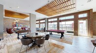 Located at 955 North Orlando Avenue in Maitland, Florida, this generously sized amenity space offers a dynamic blend of modern industrial design and comfortable social living. The main open-concept area serves as a game and entertainment hub, highlighted by a striking slatted wood ceiling detail that adds warmth and architectural interest overhead. Exposed brick accent walls contrast beautifully with light wood-plank flooring and grey shiplap paneling, creating a layered, eclectic aesthetic throughout. The space is well-equipped for recreation, featuring a full-size pool table with pendant lighting above, a foosball table, and a dedicated poker/card table with seating — ideal for social gatherings. A separate lounge area showcases curated mid-century modern furnishings, including leather egg chairs, a geometric glass coffee table, and a bold sputnik chandelier, all set against an artfully decorated shiplap feature wall. A cozy TV lounge with exposed brick and whitewashed flooring adds a relaxed nook for unwinding. Two bathrooms provide convenient facilities for guests. The expansive balcony runs the length of the space, offering open-air seating with panoramic views of the Maitland skyline and surrounding community, bathed in abundant Florida sunshine.