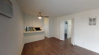 This single-floor residence offers a well-organized layout with distinct living, kitchen, sleeping, and bathroom areas, all unified by rich, dark hardwood-style flooring throughout. The open living area is generously sized, bathed in natural light from large sliding glass doors that lead to an outdoor patio enclosed by a decorative lattice screen. A ceiling fan overhead provides comfort, while a striking full-height brick fireplace with glass doors serves as a standout focal point in the room, adding warmth and character to the otherwise clean, neutral palette.

The kitchen is efficiently designed with white cabinetry, light countertops, and stainless steel appliances including a refrigerator, range, and over-the-range microwave. A pass-through counter connects the kitchen to the living space, enhancing the open feel. A window in the kitchen allows natural light to filter in.

The bedroom is comfortably proportioned, featuring a wide reach-in closet and a horizontal window that brings in additional natural light. The bathroom includes a white vanity with ample drawer storage, a large mirror, and a separate tub-shower combination with a sliding glass enclosure. White walls and clean finishes maintain a bright, consistent aesthetic throughout the space.