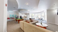 This well-appointed community lounge and recreation room offers a thoughtfully designed, open-plan layout filled with natural light and a lively, social atmosphere. The space is defined by its distinctive sculptural ceiling design, featuring large, sweeping curved recesses that add architectural character and visual depth throughout.

The lounge area anchors the room with a generously sized sectional sofa and accent chairs, complemented by colorful throw pillows and a clean round coffee table. A full-size billiards table and foosball table provide ample entertainment options. Vibrant, large-format artwork lines the walls, adding a pop of color and personality to the neutral palette.

A curved kitchenette bar serves as a central gathering point, complete with dark granite countertops, white cabinetry, a multicolored mosaic tile backsplash, pendant lighting, bar stools, stainless steel appliances, and a full refrigerator — ideal for hosting and communal use.

A sun-drenched windowed alcove with floor-to-ceiling glass panels wraps around a corner of the space, offering panoramic views of the resort-style pool courtyard and surrounding landscaped grounds. High-top bistro tables and modern seating in this bay area create an inviting spot for casual conversation. Patterned carpet flooring ties the entire space together, and glass-paneled doors hint at an adjacent fitness facility.