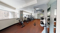 This fitness center is a comprehensively outfitted facility designed to support a wide range of workout routines. The space is laid out on a single floor with a generous, open floor plan that accommodates multiple exercise zones without feeling cramped. Underfoot, a durable patterned carpet in deep red and orange tones lines the entire workout area, providing both comfort and visual warmth throughout the space.

The cardio section is particularly well-stocked, featuring a row of elliptical trainers, a stair climber, and a spin bike, all positioned along a windowed wall that allows ample natural light to pour in and offers views of the surrounding exterior. This window-lined wall creates an open, airy atmosphere that enhances the workout environment.

A full-length mirror runs along one side of the room, visually expanding the space and allowing users to monitor their form during exercise. The functional training area is equipped with a TRX suspension system and an overhead cable machine, catering to bodyweight and resistance-based workouts. A dedicated free weights zone includes a dumbbell rack and an adjustable weight bench, while a rowing machine rounds out the cardiovascular offerings.

The ceiling features recessed lighting and structural coffers that add architectural character to the space. Tiled columns punctuate the room, contributing to its clean, modern aesthetic. The overall design is practical, well-lit, and thoughtfully arranged to maximize usability.