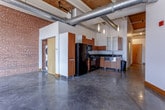 This single-floor loft showcases a compelling industrial aesthetic rooted in its original architectural character. Throughout the space, polished concrete floors reflect natural light, while exposed red brick walls add warmth and texture. Wood-beamed ceilings and prominently routed steel ductwork reinforce the raw, converted-building character that defines the overall ambiance.

The open main living area is generously proportioned, with soaring ceilings that enhance the sense of volume. Large black-framed windows allow abundant natural light to pour across the concrete floors. A stacked washer and dryer unit is conveniently integrated near the main living space.

The kitchen is well-appointed with warm wood cabinetry, dark granite countertops, a stainless steel sink, and a full suite of black appliances including a refrigerator, range, microwave, and dishwasher. Additional rooms, separated by modern framed doorways, offer flexible use as bedrooms or private workspaces.

Two bathrooms serve the unit: one features a vessel-style sink with a wood vanity cabinet and mirrored medicine cabinet, while the other includes a soaking tub with dark tile surround, a glass vessel sink atop a floating vanity, and polished concrete flooring. Together, these elements create a cohesive, urban-industrial atmosphere.