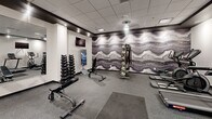 Located at 612 Sixth Street in Rapid City, South Dakota, this dedicated exercise room offers a comprehensive fitness environment within a compact, efficiently designed single-floor layout. The space is finished with a clean, modern aesthetic featuring crisp white walls, dark trim accents, and a drop-panel ceiling equipped with recessed and pendant lighting that keeps the room bright and energizing.

The flooring throughout is a durable, speckled gray rubberized surface, ideal for fitness use and easy maintenance. Cardio equipment is well-represented, with multiple treadmills and a stationary bike positioned along one wall, each accompanied by wall-mounted flat-screen televisions for entertainment during workouts. A multi-function cable machine with a weight bench anchors the strength-training area, while a full dumbbell rack with an adjustable bench rounds out the free-weight section.

A standout design element is the bold, floor-to-ceiling marble-effect accent wall in rich tones of gray, purple, and gold, lending the space a sophisticated and visually striking focal point. A mirrored wall section near the entrance enhances the sense of openness. The room is accessed through a glass-paneled door, adding a sleek, professional feel. Overall, this fitness facility is thoughtfully appointed to support a complete workout experience.