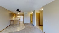 This single-floor residence offers a practical and well-organized open-concept layout that seamlessly connects the main living areas. The kitchen is equipped with warm honey-toned wood cabinetry, a white appliance suite including a refrigerator and range, and a peninsula with a tile-style countertop that provides additional prep space and informal seating. A ceiling fan with a built-in light fixture is centrally positioned, ensuring good air circulation throughout the shared living and kitchen space. The flooring transitions naturally from tile-style in the kitchen to plush carpeting in the living area, adding warmth and comfort underfoot. Neutral gray walls and a textured ceiling contribute to a clean, understated aesthetic throughout. A hallway extends from the main living space, leading to multiple doors that access private rooms, offering a clear separation between communal and personal areas. Recessed ceiling-mounted light fixtures provide consistent, functional illumination throughout the corridor and adjoining spaces. The overall layout is straightforward and efficient, making effective use of the available footprint while maintaining an open and airy feel across the main living zone.
