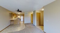 This single-floor residence offers a practical and well-organized open-concept layout that seamlessly connects the main living areas. The kitchen is equipped with warm honey-toned wood cabinetry, a white appliance suite including a refrigerator and range, and a peninsula with a tile-style countertop that provides additional prep space and informal seating. A ceiling fan with a built-in light fixture is centrally positioned, ensuring good air circulation throughout the shared living and kitchen space. The flooring transitions naturally from tile-style in the kitchen to plush carpeting in the living area, adding warmth and comfort underfoot. Neutral gray walls and a textured ceiling contribute to a clean, understated aesthetic throughout. A hallway extends from the main living space, leading to multiple doors that access private rooms, offering a clear separation between communal and personal areas. Recessed ceiling-mounted light fixtures provide consistent, functional illumination throughout the corridor and adjoining spaces. The overall layout is straightforward and efficient, making effective use of the available footprint while maintaining an open and airy feel across the main living zone.