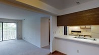 This single-floor space showcases a practical and open layout designed to maximize functionality. The living area features neutral-toned carpet, clean white walls, and a ceiling with a subtle recessed detail that adds architectural interest. A wide open archway connects the main living space to an adjoining room, enhancing the sense of flow and continuity throughout.

The kitchen is positioned adjacent to the living area and features a pass-through counter with a white surface, creating a natural divide while maintaining visual openness between spaces. A dark wood-paneled overhead cabinet adds a warm contrast to the otherwise bright, neutral palette. The kitchen is equipped with a white electric range and a stainless sink, visible through the pass-through opening. Under-cabinet lighting illuminates the workspace effectively.

The transition between the carpeted living area and the hard-surface flooring near the kitchen adds subtle visual texture. The overall ambiance of the space is clean, understated, and functional, with a neutral color scheme that provides a versatile backdrop suited to a variety of interior styles.