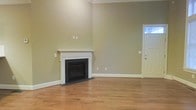 This 3-floor home presents a well-maintained interior with classic design elements. The main living area is immediately welcoming, featuring warm hardwood floors that extend throughout the space. A traditional fireplace with a crisp white surround and mantel serves as a natural focal point, adding both character and functionality to the room. The walls are finished in soft, neutral sage and warm greige tones, creating a calm and versatile backdrop suitable for a variety of interior styles.

Crown molding runs along the upper perimeter of the living room, lending a refined architectural detail that elevates the overall aesthetic. The ceilings are notably generous in height, contributing to an open and airy feel. A paneled front door with a transom window allows natural light to filter into the entryway, brightening the space near the entrance.

White baseboards provide clean contrast against the hardwood floors, maintaining a cohesive and polished finish throughout. The open layout of the main level offers flexible furniture arrangement options. Spanning three floors in total, this home provides ample vertical space and room distribution for comfortable everyday living.
