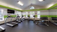 This fitness center is designed to offer a comprehensive workout environment within a single, open-plan floor. The space is immediately distinguished by its vaulted ceiling, which rises to a peak accented by a arched transom window, flooding the interior with natural light. A ceiling fan aids in air circulation throughout the room. Bold horizontal green and grey striping along the walls gives the space a vibrant, energetic aesthetic that is both modern and motivating.

The flooring consists of durable rubber matting throughout the main workout area, providing cushioning and grip suited for a range of exercise activities. Large windows fitted with blinds line multiple walls, offering views of the surrounding greenery while maintaining a bright, airy atmosphere.

The cardio section is well-appointed, featuring multiple treadmills, elliptical machines, a stair climber, and both upright and recumbent stationary bikes. A wall-mounted flat-screen television is positioned for easy viewing during cardio sessions. The strength training area includes a multi-station cable and weight machine, a free weights rack stocked with dumbbells, and a weight bench. A full-length mirror along one wall enhances the sense of space and allows for form checking during workouts. Additional fitness accessories such as medicine balls and a kettlebell are also available. The space connects to an adjacent tiled entryway, providing a seamless transition into the facility.