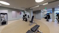 The Bishops fitness center is a comprehensive, single-floor facility designed to support a wide range of workout routines. The space is organized into distinct functional zones, each thoughtfully equipped and finished with a durable speckled rubber flooring that flows seamlessly throughout.

The cardio zone features a lineup of modern machines including ellipticals, stair climbers, and upright bikes, each equipped with personal display screens. Multiple wall-mounted flat-screen televisions provide additional entertainment, while natural light filters in through several windows to create an energizing atmosphere. Circular recessed skylights in the ceiling add a distinctive architectural touch and further brighten the space.

The strength training area is outfitted with a comprehensive selection of cable and resistance machines, free-weight benches, and a well-stocked dumbbell rack. Large floor-to-ceiling mirrors line the walls, enhancing the sense of openness and allowing users to monitor their form. Soft, neutral grey walls and clean recessed lighting contribute to a polished, professional environment.

A dedicated spin studio houses multiple Peloton-style cycling bikes with integrated screens, complemented by medicine balls, stability balls, a punching bag, and ample storage for yoga mats and accessories — making it ideal for group classes or individual cross-training. The facility is climate-controlled throughout and finished with a clean, modern aesthetic.