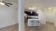 Located in the Wolf Ranch community of Georgetown, Texas, this two-story residence showcases a thoughtfully designed interior with modern finishes and an open, airy feel. The main living area flows seamlessly into the kitchen, which serves as the centerpiece of the space. Rich dark cabinetry creates a striking contrast against the crisp white subway tile backsplash and the smooth white countertops, lending the kitchen a sleek, contemporary character. Stainless steel appliances, including a French door refrigerator, a built-in microwave, and a range, add a polished, functional touch. A curved peninsula with bar-height seating and modern metal stools provides an inviting spot for casual dining or entertaining. Pendant lights and a track lighting system illuminate the kitchen, while the adjoining living area features a ceiling fan with a light fixture. Light-toned hardwood-style flooring runs throughout the main level, enhancing the open and cohesive feel of the space. Neutral walls keep the palette fresh and versatile, allowing the bold cabinetry and fixtures to make a strong visual statement. Spanning two floors, this residence offers well-defined living spaces with a modern aesthetic suited for comfortable everyday living.