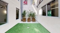 Pet Wag is a single-story pet care facility centered around a well-appointed outdoor courtyard designed with both pets and their owners in mind. The exterior façade features a smooth stucco finish with large windows that allow natural light to filter into the interior spaces while providing visual connectivity between the indoor and outdoor areas. Through the windows, interior rooms are visible with wall-mounted lighting and potted plants, suggesting a clean and carefully maintained environment.

The courtyard serves as the focal point of the space, finished with vibrant artificial turf that provides a safe, easy-to-clean surface for animals. Decorative pea gravel borders line the perimeter, complementing large square metallic planters that house lush bamboo plants and ornamental grasses, adding a natural, calming aesthetic. The vertical scale of the building walls surrounding the courtyard creates an open-air feel while maintaining an enclosed, secure environment.

Practical details include drainage grates embedded in the turf, a hose reel station, and a utility door for staff access, reflecting the facility's operational functionality. Colorful artwork displayed on upper-level windows adds a playful, welcoming touch. Overall, the space balances aesthetic appeal with practical design tailored to pet care.