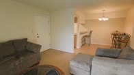 This single-floor space offers a practical and welcoming layout designed for everyday comfort. The living area serves as the heart of the home, featuring light tile flooring and neutral-toned walls that create a bright, airy atmosphere. Soft seating and an accent rug anchor the space, providing a cozy gathering area. A partial wall with a pass-through opening connects the living room to the kitchen and dining area, promoting an open-concept feel while maintaining a sense of definition between spaces. The dining area is highlighted by an elegant chandelier overhead, lending a touch of classic charm, while a raised breakfast bar with bar stools offers a casual spot for dining or conversation. The kitchen, visible beyond the pass-through, features warm wood-toned cabinetry and appears well-appointed for daily use. The front entry door is conveniently accessible from the living area, adding to the efficient flow of the floor plan. Throughout the space, clean white baseboards and trim provide a crisp finish. The overall ambiance is calm, functional, and move-in ready, making it a comfortable and straightforward living environment.