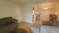 This single-floor space offers a practical and welcoming layout designed for everyday comfort. The living area serves as the heart of the home, featuring light tile flooring and neutral-toned walls that create a bright, airy atmosphere. Soft seating and an accent rug anchor the space, providing a cozy gathering area. A partial wall with a pass-through opening connects the living room to the kitchen and dining area, promoting an open-concept feel while maintaining a sense of definition between spaces. The dining area is highlighted by an elegant chandelier overhead, lending a touch of classic charm, while a raised breakfast bar with bar stools offers a casual spot for dining or conversation. The kitchen, visible beyond the pass-through, features warm wood-toned cabinetry and appears well-appointed for daily use. The front entry door is conveniently accessible from the living area, adding to the efficient flow of the floor plan. Throughout the space, clean white baseboards and trim provide a crisp finish. The overall ambiance is calm, functional, and move-in ready, making it a comfortable and straightforward living environment.