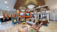 Located on N High Street in Columbus, Ohio, Luxe Belle is a thoughtfully designed amenity floor that combines bold aesthetics with practical, community-focused spaces. The main lounge is an expansive gathering area anchored by a striking orange accent wall, a fireplace, and dual mounted flat-screen TVs. Plush seating arrangements — including tufted sofas, wingback chairs, and leather accent chairs — are layered over vibrant patterned rugs, creating an inviting and eclectic atmosphere. Overhead, dramatic drum pendant lighting and a warm wood-paneled ceiling add architectural depth to the space. Light wood-look flooring with hexagonal tile inlays flows throughout, lending continuity across zones. A popcorn machine and bar-height seating add a casual, social element near large floor-to-ceiling windows that open views to an outdoor patio area. A dedicated coffee and beverage bar features dark wood cabinetry, a beverage cooler, and a full coffee station set against a patterned tile backsplash, framed by a white brick partition with pendant lighting. A secondary lounge nook, marked by bold "BELLE" wall art and a Columbus-inspired mural, sits adjacent to the fully equipped fitness center, visible through glass partitions. A private tanning room rounds out the amenity offerings, providing a complete lifestyle-focused experience.