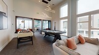 Located at 3986 Wilshire Blvd in Los Angeles, this single-floor amenity space is designed as a contemporary game and social lounge. The room immediately impresses with its soaring double-height ceilings, which are accentuated by dramatic pendant lighting fixtures with geometric, circular designs that cascade from above, serving as a striking focal point. Large floor-to-ceiling windows line the walls, flooding the interior with abundant natural light while offering views of the surrounding urban environment.

The space is centered around a full-sized pool table with a sleek dark wood frame and light felt surface, complemented by a foosball table nearby, making it an ideal setting for recreational gatherings. Comfortable, neutral-toned sofas with warm accent pillows are thoughtfully arranged around a bold black-and-gold striped area rug, creating a relaxed lounge area. A wall-mounted flat-screen television is positioned above a light wood credenza, adding a media component to the space.

Decorative wall murals and large-scale botanical artwork lend a vibrant, artistic character to the white walls. An adjacent area, visible through dark wood-framed openings, offers additional lounge seating with outdoor views, enhancing the overall sense of openness and connectivity throughout the space.