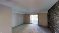 This single-floor space features an open living area finished with wall-to-wall carpeting in a neutral tone, contributing to a calm and understated ambiance. The room is generously proportioned, providing ample space for a variety of furniture arrangements. Smooth, flat ceilings with subtle crown molding detailing run throughout, lending a clean and classic architectural character to the interior. The walls are finished in a soft, warm beige, creating a neutral backdrop that suits a wide range of interior styles. A prominent sliding glass door serves as the room's focal point, allowing natural light to filter in and offering a direct view of the outdoor greenery beyond. Vertical blinds accompany the sliding door, providing flexible light and privacy control. The transition between the carpeted interior and the sliding door threshold is clean and well-defined. Multiple electrical outlets are positioned at regular intervals along the walls, reflecting practical and functional planning throughout the space. Overall, the room presents a bright, versatile, and move-in-ready environment with a simple, comfortable aesthetic suited to everyday living.