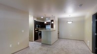 This single-floor residence offers a thoughtfully designed open-concept layout that seamlessly connects the kitchen and living areas. The kitchen is a standout feature, outfitted with dark espresso cabinetry that provides a rich contrast against the neutral-toned walls. Stainless steel appliances, including a refrigerator, microwave, and range, bring a modern touch to the space. A peninsula breakfast bar extends from the kitchen, offering additional prep space and a casual seating option while maintaining a clear visual connection to the rest of the unit. Pendant lighting hangs above both the kitchen and the dining area, adding warmth and definition to each zone. The living area is finished with plush, light-toned carpeting that contributes to a comfortable and inviting ambiance. A decorative pendant chandelier anchors the dining space, adding a subtle design element. The overall color palette—soft beiges and warm neutrals throughout—creates a cohesive and calming atmosphere. The layout is efficient and functional, making excellent use of the available footprint while maintaining an open, airy feel throughout the main living spaces.