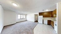 This single-floor space features an open-concept layout that seamlessly connects the living area to the kitchen. The living area is finished with wall-to-wall neutral carpeting, white walls, and a textured ceiling, creating a clean and functional environment. A window along the exterior wall allows natural light to filter into the space, brightening the interior. The transition from carpet to tile flooring marks the boundary between the living space and the kitchen area. The kitchen is equipped with white appliances, including a full-size refrigerator and dishwasher, paired with medium-toned wood cabinetry that provides ample storage. The tile flooring in the kitchen offers a practical, easy-to-clean surface. A hallway visible from the main living area leads to additional rooms within the unit. Flush-mount ceiling light fixtures provide consistent overhead lighting throughout. The overall layout is straightforward and efficient, with a neutral color palette that offers a versatile backdrop suitable for a variety of furnishing styles.