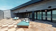Located at 500 W Broad St in Columbus, Ohio, this contemporary single-story property makes a strong architectural statement with its sleek, modern design. The exterior is clad in dark horizontal siding panels that lend a sophisticated, urban aesthetic, complemented by clean lines and a low-profile roofline that emphasizes a sense of understated refinement.

The standout feature of this space is its expansive outdoor deck, finished with rich natural wood tile flooring laid in a classic pattern that adds warmth and texture to the otherwise bold, dark palette of the building. The deck is thoughtfully appointed with modern outdoor lounge furniture, including cushioned chaise lounges in teal and neutral tones, creating an inviting atmosphere ideal for relaxation or casual gatherings.

Large floor-to-ceiling glass sliding doors connect the outdoor deck directly to the interior, allowing for an effortless indoor-outdoor flow while flooding the adjacent interior spaces with natural light. Exterior wall-mounted lighting fixtures add both functionality and style to the façade.

The overall ambiance of the property is one of contemporary sophistication — a harmonious blend of natural materials and modern architectural elements that creates a polished, functional environment well-suited to its Columbus address.