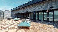 Located at 500 W Broad St in Columbus, Ohio, this contemporary single-story property makes a strong architectural statement with its sleek, modern design. The exterior is clad in dark horizontal siding panels that lend a sophisticated, urban aesthetic, complemented by clean lines and a low-profile roofline that emphasizes a sense of understated refinement.

The standout feature of this space is its expansive outdoor deck, finished with rich natural wood tile flooring laid in a classic pattern that adds warmth and texture to the otherwise bold, dark palette of the building. The deck is thoughtfully appointed with modern outdoor lounge furniture, including cushioned chaise lounges in teal and neutral tones, creating an inviting atmosphere ideal for relaxation or casual gatherings.

Large floor-to-ceiling glass sliding doors connect the outdoor deck directly to the interior, allowing for an effortless indoor-outdoor flow while flooding the adjacent interior spaces with natural light. Exterior wall-mounted lighting fixtures add both functionality and style to the façade.

The overall ambiance of the property is one of contemporary sophistication — a harmonious blend of natural materials and modern architectural elements that creates a polished, functional environment well-suited to its Columbus address.