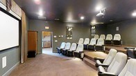 Located at 560 New Hope Dr in Goldsboro, North Carolina, this single-floor space is purpose-built for an immersive cinematic experience. The centerpiece of the space is a dedicated screening room finished in deep, dark tones that create a true theater ambiance. Rows of plush, black-and-cream leather recliner-style seats are arranged on a tiered, raised platform, ensuring excellent sightlines from every position. A ceiling-mounted projector anchors the room as the primary viewing system, complemented by recessed lighting that allows for adjustable atmospheric control. Framed artwork on the walls adds a touch of character to the otherwise sleek, cinema-inspired interior. Wall-mounted audio and control panels suggest a fully integrated audiovisual setup. Adjacent to the main screening area is a well-appointed kitchenette, featuring cabinetry, countertops, and a sink — providing convenient access to refreshments without leaving the space. The carpeted flooring throughout adds warmth and contributes to the room's acoustic qualities. Overall, the space delivers a polished, professional theater environment well-suited for private screenings, presentations, or entertainment gatherings.