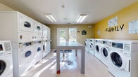 This shared laundry facility is a single-floor space designed for efficient, community use. The room is organized into clearly defined functional zones, with rows of front-loading washing machines lining one wall beneath a bold yellow accent wall bearing the word "FAMILY," creating a warm and welcoming atmosphere. Opposite, stacked dryer units maximize the use of vertical space, providing ample drying capacity for multiple users simultaneously. A centrally positioned folding table offers a convenient surface for sorting and folding laundry, keeping the workflow organized. The light-toned tile flooring is easy to maintain and reflects the bright fluorescent overhead lighting, ensuring the space is well-lit throughout. A glazed exterior door allows natural light to filter into the room, adding to the overall brightness and providing a view of the surrounding outdoors. A seating area near the entrance includes a chair for those waiting, alongside a wooden interior door that leads to an adjacent space. Instructional and service signage is posted throughout, offering guidance on machine use and laundry tips. The combination of generous machine capacity, practical layout, and cheerful design details makes this a functional and comfortable communal laundry space.