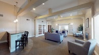 This single-floor space is thoughtfully designed as a multi-functional gathering facility, incorporating several distinct areas that flow naturally into one another. The architecture is characterized by vaulted ceilings with decorative beam detailing finished in warm tan tones, recessed lighting throughout, and neutral wall colors that contribute to a calm, welcoming atmosphere.

The main lounge area anchors the space, featuring plush gray sofas and armchairs arranged around a stone-surround fireplace, flanked by windows that allow natural light to filter in through slatted blinds. Decorative plants and blue accent pillows add warmth and color to the seating arrangement. French doors with glass panel inserts connect this area to adjacent spaces, enhancing the open feel.

A dedicated meeting or activity room accommodates multiple tables and chairs, suitable for group gatherings or events. Large windows along the walls provide generous natural light and views to the outdoors, while vaulted ceilings lend an airy quality to the room.

A kitchenette area features a curved dark granite countertop bar with bar seating, pendant lighting overhead, and a separate counter with a sink and cabinetry, making it well-suited for catering or refreshments during events. Tile flooring grounds this functional zone. Together, these areas create a versatile and comfortable community environment.