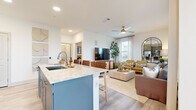 This thoughtfully designed 2-bedroom, 2-bathroom residence spans a single floor, offering an efficient and comfortable open-concept layout. The combined kitchen and living area serves as the heart of the home, anchored by a large kitchen island with a marble-style countertop, undermount sink, and seating for multiple bar stools — ideal for casual dining and entertaining. The kitchen features gray shaker cabinetry, a subway tile backsplash, and a full suite of stainless steel appliances. Light hardwood-style flooring runs throughout the main living areas, complementing the bright, white walls and recessed lighting overhead. The living room is generously sized, bathed in natural light through multiple windows, and fitted with a ceiling fan. The primary bedroom offers plush carpeting, a ceiling fan, a dedicated en-suite bathroom, and direct access to a spacious walk-in closet with built-in shelving. The second bedroom is equally well-sized with comfortable carpeting. Both bathrooms are finished with gray shaker vanities, marble-style countertops, and framed mirrors. A utility room and two closets provide ample storage throughout. The overall ambiance is modern, clean, and welcoming.