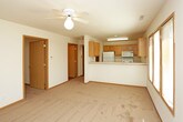 This single-floor residence offers a practical and functional layout connecting several distinct living areas. The galley-style kitchen is equipped with warm oak cabinetry on both sides, white appliances including a range with oven and built-in microwave, light-toned countertops, a stainless steel sink, and durable wood-look flooring. A peninsula counter separates the kitchen from the adjoining living area, creating an open-concept feel between the two spaces.

The living area is generously sized, featuring wall-to-wall carpet in a neutral tan tone, a ceiling fan with a light fixture, and a row of windows fitted with horizontal blinds that allow natural light to fill the room. The carpet continues into the bedrooms, one of which features a pair of windows providing additional natural light, while another includes its own ceiling fan. Both rooms offer clean, neutral walls and ample space.

The bathroom is well-proportioned, featuring wood-look flooring consistent with the kitchen, a full-size soaking tub with a shower, a toilet, and a wood-toned vanity with countertop. Throughout the unit, wood-trimmed doors and windows, neutral wall tones, and consistent flooring materials create a cohesive and comfortable aesthetic.