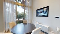 This refined single-floor space exudes a polished, contemporary aesthetic that seamlessly blends comfort with sophistication. The interior is anchored by generous floor-to-ceiling windows dressed in sheer, light-filtering curtains that bathe the space in abundant natural light while framing lush tropical palm views beyond — creating a seamless connection between indoors and the surrounding landscape.

A standout dining or meeting area features a sleek, dark circular table paired with plush upholstered chairs in soft neutral tones, resting atop a boldly patterned area rug that adds visual interest to the refined palette. The furniture selection reflects a careful attention to detail, balancing form and function.

On the opposite wall, a large wall-mounted flat-screen display is recessed for a clean, streamlined look, accompanied by a contemporary low-profile credenza in light stone or lacquered finish, offering both storage and surface space. Overhead, a cluster of Edison-style pendant lights suspended at varying heights adds warmth and a touch of modern industrial character to the otherwise serene, neutral-toned interior.

The overall ambiance is bright, airy, and inviting — ideal for both productive gatherings and relaxed conversation — with a design language that speaks to modern elegance and thoughtful spatial planning.