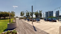 Located at 100 Park Ave in Fort Lee, NJ, this thoughtfully designed outdoor amenity space offers a comprehensive range of recreational and leisure facilities set against the striking backdrop of a glass-curtain high-rise tower. The grounds are anchored by a resort-style swimming pool surrounded by a spacious tiled deck, pergola-shaded lounge chairs, and blue umbrellas, creating an inviting sunbathing and relaxation area. Adjacent to the pool, a dedicated terrace features fire pit tables and plush blue lounge seating strung with overhead bistro lighting, ideal for evening gatherings. Manicured green lawns and curved garden beds with ornamental shrubs and trees add lush natural character throughout the space. A separate terrace zone incorporates large cylindrical planters and additional seating areas that open to panoramic urban skyline views. Families are well served by a vibrant children's play area with colorful rubber surfacing, while sports enthusiasts can take advantage of a full outdoor basketball court. Landscaped pathways lined with ornamental grasses and mature trees connect each zone seamlessly. The overall ambiance balances active recreation with tranquil green space, all set within a dynamic urban environment with open sky and broad city views.