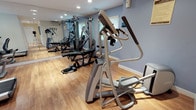 This dedicated fitness center is thoughtfully designed to support a comprehensive workout experience. The space is finished with warm hardwood flooring throughout, providing a clean and durable surface ideal for exercise. Recessed lighting illuminates the room evenly, complemented by natural light streaming in through multiple windows fitted with slatted blinds.

The gym is outfitted with a well-rounded selection of equipment, including a multi-station cable and weight machine, a stationary recumbent bike, a stair climber, an elliptical trainer, a treadmill, and a flat workout bench. A wall-mounted flat-screen television is positioned for viewing during cardio sessions, adding convenience to longer workouts.

Floor-to-ceiling mirrors line one wall, visually expanding the space and allowing users to monitor their form during exercise. A split-system air conditioning unit ensures climate control year-round, maintaining a comfortable environment regardless of the season. A wall-mounted hand sanitizer dispenser reflects attention to hygiene standards.

A connecting corridor with recessed lighting and additional doors leads to adjoining rooms, suggesting further amenities nearby. The overall ambiance is clean, functional, and professional, making this fitness center a practical and well-appointed facility for a variety of training needs.