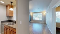 This single-floor space offers a well-organized layout that balances open living areas with functional private spaces. The main living area is finished with light wood-style hardwood flooring and neutral gray walls, creating a bright and welcoming atmosphere. Large windows along the exterior wall allow generous natural light to fill the room, with views of greenery outside. Wall sconces and a pendant chandelier provide additional ambient lighting throughout.

The kitchen is efficiently designed, featuring warm-toned wooden cabinetry, gray subway tile backsplash, solid countertops, and a full suite of stainless steel appliances including a refrigerator, electric range, and over-the-range microwave. A pass-through opening connects the kitchen visually to the main living area. In-unit laundry is also accessible from the main living space.

The bedroom is carpeted with a neutral tone and includes a built-in closet with a hanging rod for ample storage. A tray ceiling detail with a contrasting accent color adds architectural interest to the room.

The bathroom features large-format tile flooring in a warm beige tone, a soaking tub, toilet, and clean white walls, presenting a simple and functional design. A wall-mounted grab bar adds a practical touch. Overall, the space maintains a clean, contemporary aesthetic throughout.