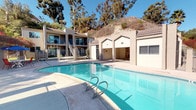 Located at 501-567 Canyon in Oceanside, this inviting outdoor pool area offers a well-maintained communal retreat surrounded by lush natural scenery. The centerpiece is a generously sized swimming pool with crystal-clear blue water, accessible via stainless steel entry rails and framed by a wide, smooth concrete sundeck. The deck provides ample space for lounging and outdoor dining, with patio furniture and umbrella-shaded seating areas positioned poolside for relaxed gatherings under the Southern California sun.

A dedicated single-story amenity building anchors one side of the pool area, featuring arched architectural detailing and tiled roofing that complement the warm, neutral stucco exterior. The structure houses utility and support facilities for the pool area. Secure wrought-iron fencing encloses the entire pool courtyard, offering both safety and a defined sense of place.

Tall eucalyptus trees and palm trees rise dramatically above the property, framing views of the natural hillside that backs the complex and lending a serene, resort-like atmosphere. The multi-story residential building overlooks the pool area with private balconies, enhancing the sense of community. Under sunny blue skies typical of coastal Southern California, this pool area delivers a bright, open, and welcoming outdoor environment year-round.