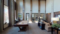This single-floor community space is defined by its dramatic, soaring ceilings that lend an open, grand atmosphere throughout. Prominent wood and stone-clad columns rise from floor to ceiling, anchoring the expansive main lounge area and adding a sense of architectural refinement. A patterned carpet with a leaf motif runs across the main floor, complementing the warm, earthy tones of the interior palette.

The central gathering area features a full-size billiards table as a focal point, surrounded by comfortable lounge seating arranged around a flat-screen television mounted on a textured wood-paneled wall flanked by stacked stone columns. A bar-height counter with industrial-style stools provides an additional social hub. Tall floor lamps and pendant lighting contribute to the warm, inviting ambiance.

Multiple glass-panel doors open to adjacent rooms, including a dining and conference area, as well as an exterior patio, allowing abundant natural light to filter throughout the space.

The kitchen area is well-appointed with dark espresso cabinetry, granite countertops, a mosaic tile backsplash, stainless steel appliances including a large built-in refrigerator and dishwasher, and a frosted-glass pantry door—offering a polished, functional space suited for gatherings and events.