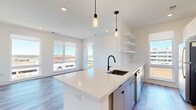 This contemporary single-floor residence showcases a clean, modern design highlighted by light wood-look flooring, crisp white walls, and black hardware accents throughout. The open-concept kitchen is a standout feature, centered around a large waterfall-edge quartz island with an undermount sink and matte black pull-down faucet. Sleek flat-front cabinetry in white and gray pairs with stainless steel appliances, including a range, built-in microwave, and refrigerator, while a classic white subway tile backsplash and open floating shelves complete the look. Pendant lighting illuminates the island, which flows seamlessly into the living and dining area, where large windows frame urban skyline views and flood the space with natural light. A private balcony, accessible through a glass-paneled door, extends the living space outdoors. The bedroom features a ceiling fan, generous windows with city views, and direct access to a walk-in closet equipped with wire shelving. The bathroom offers marble-look tile flooring, a white vanity with black hardware, and a backlit mirror. Recessed lighting and roller shades are found throughout, contributing to the residence's polished, move-in-ready aesthetic.