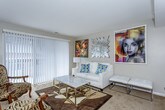 This single-floor residence offers a thoughtfully arranged layout that balances comfort and artistic character throughout. The living room serves as the heart of the home, showcasing bold, large-format artwork adorning warm neutral walls, complemented by plush carpeting underfoot. A marble-topped coffee table, a white sofa, and eclectic accent chairs create an inviting yet stylish atmosphere. Sliding glass doors and surrounding windows flood the space with natural light while providing access to an outdoor area, enhancing the open, airy feel of the room. A separate dining area with a glass table sits adjacent, completing the communal living zone.

The kitchen is well-appointed with dark cabinetry, granite-style countertops, a white tile mosaic backsplash, and a full suite of stainless steel appliances, including a range, over-the-range microwave, and double sink — offering both functionality and a sleek, modern aesthetic.

Two carpeted bedrooms provide restful retreats, each decorated with curated artwork. One bedroom features a bold dark wood dresser and bed frame, while the other is finished in softer, neutral tones with understated wall art and warm lighting.

The bathroom is clean and practical, featuring tile flooring, a tub-shower combination with sliding glass doors, a vanity with ample counter space, and neutral finishes throughout.