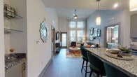 This thoughtfully designed single-story residence showcases a seamless open-concept layout connecting the kitchen, dining, and living areas. The living room stands out with dramatically high ceilings and tall windows that draw in abundant natural light, complemented by a ceiling fan and wood-style flooring throughout. A glass-paneled door provides access to the outdoors.

The kitchen is well-equipped with stainless steel appliances, including a refrigerator, range, microwave, and dishwasher. Gray shaker-style cabinetry pairs with granite countertops and a white subway tile backsplash. A granite-topped island with seating serves as both a functional workspace and casual dining area, illuminated by pendant lighting overhead.

The bedrooms feature plush carpeting, ceiling fans, and large windows offering outdoor views. Each room provides a comfortable, well-lit retreat with ample space.

The bathroom is finished with marble-style tile flooring, a tiled soaking tub and shower combination, a granite vanity countertop with an undermount sink, and gray cabinetry. An adjoining hallway includes built-in shelving and closet storage, adding practical organization to the space.