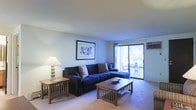 This single-floor space offers a thoughtfully arranged living area that balances comfort and functionality. The living room serves as the centerpiece, featuring plush wall-to-wall carpeting that adds warmth underfoot. A large sectional sofa in deep navy provides generous seating and anchors the room, complemented by a classic wood coffee table and coordinating end table with a soft-glow lamp that enhances the cozy ambiance. Framed artwork on the wall adds a curated, decorative touch to the neutral-toned walls. A standout feature of the living room is the sliding glass door that opens to a private balcony, allowing natural light to filter into the space and offering a seamless connection to the outdoors. The balcony, visible through the glass, is furnished with a small bistro-style table and chairs, making it a charming spot for relaxing or dining al fresco. A wall-mounted air conditioning unit near the balcony door ensures year-round climate comfort. The overall ambiance of the space is calm and inviting, with a classic design aesthetic that makes it both practical and livable.