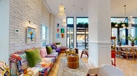 Located in the heart of Victoria, BC, the Pandora Ave Terrace Lounge is a thoughtfully designed communal space that combines eclectic style with functional amenities across a single floor. The main living and lounge area immediately impresses with soaring ceilings, exposed white brick walls, and floor-to-ceiling windows that flood the space with natural light while offering views of the surrounding urban landscape. Colorful, low-profile modular sofas, layered area rugs, brass coffee tables, and a striking arched fireplace with dark marble surround create a lively yet inviting atmosphere. Pendant lighting and curated wall art add further character throughout. The kitchen is equally well-appointed, featuring warm wood cabinetry, marble countertops, stainless steel appliances, and a decorative patterned tile floor. A bar counter with bentwood stools sits beneath an elegant arched alcove, ideal for casual gatherings. The hallway entry is refined, with a gold-framed mirror, a cane-front console, and lush greenery setting a welcoming tone. A dedicated game room provides additional entertainment space. The lounge opens directly onto a spacious outdoor terrace, furnished with plush modular seating and fire features, perfectly suited for socializing in Victoria's mild climate.