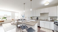 This well-designed 2-bedroom, 1-bathroom residence in Glenview, IL offers a thoughtful single-floor layout with modern finishes throughout. The open-concept kitchen and living area serves as the heart of the home, anchored by a sleek kitchen featuring crisp white shaker cabinetry, subway tile backsplash, stainless steel appliances, and a spacious island with a taupe countertop and bar seating — ideal for casual dining and entertaining. Pendant lighting adds a contemporary touch above the island. The living room receives generous natural light through large windows and features durable hardwood-style flooring, creating a bright and welcoming atmosphere. A distinctive blue accent wall carries through both the living area and the primary bedroom, adding a bold design element. The primary bedroom offers comfortable carpeting, a window with outdoor views, and a closet with built-in shelving. The bathroom is finished with warm gray walls, dark hardwood-style flooring, and a shower/tub combination. A hallway with additional closet space completes the layout. The overall aesthetic is clean and contemporary, with a neutral palette accented by modern fixtures and thoughtful design details throughout.