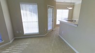 This single-floor space offers a welcoming and functional layout designed for comfortable everyday living. The open-concept living area is finished with soft, neutral-toned carpeting that adds warmth underfoot, while the walls are painted in a gentle beige tone that enhances the overall sense of brightness and calm throughout the space. Large windows dressed with white horizontal blinds allow abundant natural light to flood the interior, creating an airy and inviting atmosphere. A glass-paneled door provides access to the outdoors, further connecting the living space to the exterior and adding additional light. The living area flows seamlessly into the kitchen zone, which features a raised counter or breakfast bar with a pendant light overhead, making it an ideal spot for casual dining or entertaining. The kitchen's elevated counter provides a natural visual and functional separation between the two areas without compromising the open feel of the overall layout. The clean white trim and baseboards offer a crisp contrast to the warm wall tones, lending a polished, well-maintained appearance to the space. Overall, this single-floor layout combines practical design with a light-filled, neutral aesthetic that provides a versatile and move-in-ready environment.