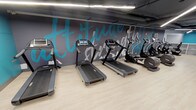 This fitness facility is thoughtfully organized across a single level, offering a comprehensive range of workout zones designed to support diverse training needs. The main cardio area features a row of treadmills and elliptical machines positioned against a striking dark grey accent wall adorned with bold teal and white motivational typography, creating an energizing atmosphere. Linear LED lighting runs throughout, illuminating light wood-style flooring that unifies the space.

The strength training zone is equipped with cable machines, a power rack with TRX suspension trainers, adjustable benches, a full dumbbell rack, kettlebells, battle ropes, and stability balls, all set against vibrant teal accent walls. A dedicated cycling studio houses stationary bikes with individual screens and features a large glass partition offering sightlines into the main gym floor, along with an exposed industrial ceiling fitted with linear lighting.

A separate functional training room with rubber flooring and a wall-mounted TV provides flexible space for group or individual workouts. A smaller activity room with a playful patterned floor, yoga mats, and soft seating offers a calm retreat for stretching or mindfulness activities. Full-length mirrors, mounted televisions, and built-in shelving complete the well-appointed facility.