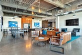 Located at 1901 East 1st Street in Santa Ana, CA, this expansive single-floor communal lounge offers a thoughtfully designed, industrial-chic environment that blends social, recreational, and lounge functions seamlessly. The open-plan layout is anchored by polished concrete floors and soaring exposed ceilings with visible ductwork, structural beams, and Edison-bulb pendant lighting that lend the space a modern, urban character.

The main living area is generously furnished with leather sofas and lounge chairs arranged atop teal accent rugs, complemented by vibrant abstract artwork adorning the walls. A full kitchen with warm wood cabinetry, stainless steel countertops, and a central island provides a functional gathering hub. A large communal dining table with ample seating sits adjacent to floor-to-ceiling windows, flooding that zone with natural light and offering views of the exterior.

Recreational amenities are a standout feature, including a foosball table, ping-pong table, billiards table, and a giant Connect Four game. A separate, more intimate lounge nook is finished with dark wood-paneled walls, built-in banquette seating, and a wall-mounted display, creating a quieter retreat within the broader space. Multiple mounted flat-screen televisions are distributed throughout, and a sliding barn door separates zones for added versatility. The overall ambiance is energetic yet comfortable, designed for both active engagement and relaxed socializing.
