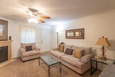 This single-floor space is thoughtfully laid out to maximize functionality and comfort throughout. The living area serves as the heart of the home, featuring warm carpet flooring, a ceiling fan with light fixture, crown molding, and a tiled fireplace flanked by a wall-mounted television. Built-in shelving along one wall provides attractive display and storage space, while a glass-top coffee table and upholstered seating complete the inviting atmosphere. A decorative mirror and framed artwork add character to the room.

Adjacent to the living area, a compact dining nook accommodates a round wooden table with chairs, accented by framed artwork above. The galley-style kitchen is efficiently designed with light wood cabinetry, dark countertops, and wood-look flooring. Black appliances, including a refrigerator, electric range, dishwasher, and built-in microwave, provide full functionality, while a charming wooden interior door adds a rustic touch.

The bedroom offers a tranquil retreat with plush carpet, a ceiling fan, crown molding, and a window dressed with rich burgundy curtains that allow natural light. A nightstand with lamp and ample door storage complete the room. Throughout the space, consistent wainscoting and neutral wall tones create a cohesive, well-finished aesthetic.