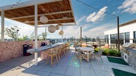Located at 645 Division St in Nashville's dynamic Gulch neighborhood, this rooftop amenity deck offers an expansive outdoor experience with panoramic views of the city skyline and surrounding mountains. The open-air terrace is anchored by a pool with shallow-entry access, lined with premium chaise lounges and shaded pergola structures ideal for relaxing under the Tennessee sun. A covered outdoor bar area features a long counter finished in pink tile, bar-height seating, and pendant globe lighting suspended from a warm wood-slatted ceiling, creating an inviting social hub. String lights and a mix of wicker and patterned accent chairs add to the relaxed yet stylish atmosphere across multiple seating vignettes. A bold, large-scale mural painted in vibrant geometric and abstract patterns adorns the central structure's exterior wall, serving as a striking visual focal point. Outdoor dining tables with eclectic seating are arranged nearby, complemented by built-in grilling stations. The deck's large-format porcelain tile flooring and corrugated metal architectural cladding give the space a modern industrial character. Greenery is woven throughout, softening the urban aesthetic. This rooftop destination seamlessly blends art, leisure, and entertainment against Nashville's iconic skyline backdrop.