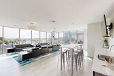 Located in Medford, MA, the City Lounge & Patios is a sophisticated rooftop amenity space designed to blend indoor comfort with outdoor living. The interior lounge is defined by dramatic floor-to-ceiling curtain-wall glazing that wraps around the perimeter, flooding the space with natural light and framing sweeping panoramic views of the Medford cityscape and surrounding treetops. Light hardwood-style flooring and crisp white ceilings contribute to the airy, contemporary aesthetic throughout. The lounge area is furnished with sleek black modular sofas arranged over a vibrant teal-accented area rug, complemented by modern drum pendant lighting. A high-top communal table with metal bar stools provides an additional seating arrangement suited for casual gatherings or collaborative use. Large sliding glass doors connect the interior to a generously sized outdoor patio finished in large-format stone tile. The patio is outfitted with multiple seating configurations, including lounge chairs, wicker armchairs, and a dining table with chairs, offering versatile options for relaxation and outdoor entertaining. Wall-mounted lighting fixtures on the surrounding building facade enhance the patio's usability. Together, the interior lounge and outdoor patio create a seamless, elevated retreat with abundant natural light and commanding views of the surrounding area.