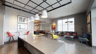 This single-floor space is a thoughtfully designed communal amenity area that combines recreation, relaxation, and social gathering in a cohesive, contemporary environment. The main lounge opens with a striking reception counter beneath an industrial-style grid ceiling fitted with pendant lighting, creating an immediate visual focal point. Colorful accent furniture — including a vibrant yellow armchair and bold orange cushions — punctuates the otherwise neutral gray palette, lending the space an energetic, modern character.

The game room features a full-size pool table set against a dramatic dark-gray backdrop, with recessed alcoves built into the walls. Each alcove is distinctly designed — one housing a flat-screen television with bar stool seating, another offering a cozy banquette with decorative brick detailing, ideal for intimate conversation or viewing.

A dedicated lounge area showcases tall wingback chairs in steel blue upholstery, framed artwork, and a colorful geometric area rug, all positioned alongside floor-to-ceiling windows that reveal panoramic urban views and connect to an outdoor terrace. A separate dining nook provides a round table with modern chairs beneath large windows, bathed in natural light with views of the surrounding cityscape. Together, these zones create a versatile, design-forward space suited for both casual socializing and quiet retreat.